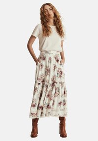 Kwiatowa spódnica maxi w kremowym kolorze z różowymi i zielonymi wzorami, z falbanowym krojem, elastycznym paskiem w talii i bocznymi kieszeniami; zestawiona z białym t-shirtem.