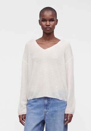 Personne aux cheveux courts portant un pull en maille blanc ample à col en V et un jean bleu clair, debout devant un fond uni clair.