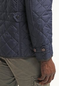 Marineblå quiltet jakke med knaplukket manchet, glat tekstur, diagonalt mønster og kontrasterende brune knapper. Oliemærkede bukser synlige nedenunder.