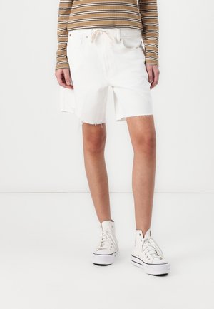 Personne portant un short en denim blanc effiloché, des baskets montantes blanches et un t-shirt à manches longues rayé marron, debout sur un fond blanc uni.