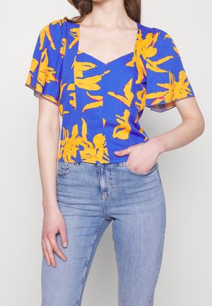 Woman wearing blue blouse with yellow floral print and puffed short sleeves, paired with light blue denim jeans.