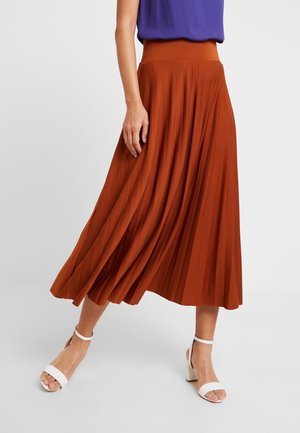 Vrouw draagt een roestoranje geplooide midi-rok, paarse top en witte hoge hakken met enkelriempje, staand met één voet naar voren.