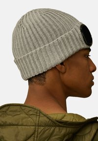 Cappello in maglia grigia con texture a costine e risvolto largo, caratterizzato da un grande accento circolare nero sul lato, indossato da una persona in giacca verde.