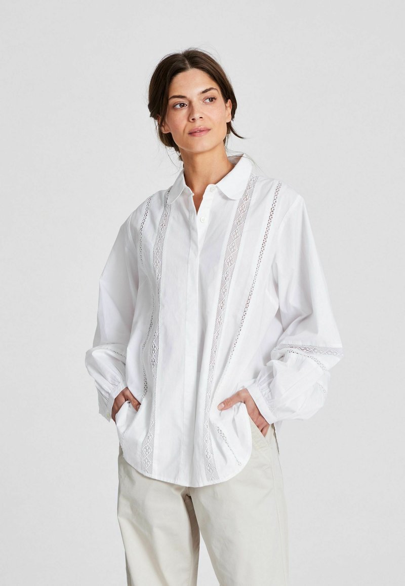Camicia bianca con colletto e maniche lunghe, caratterizzata da dettagli in pizzo decorativi lungo la parte anteriore e tasche sui lati. Materiale in cotone.