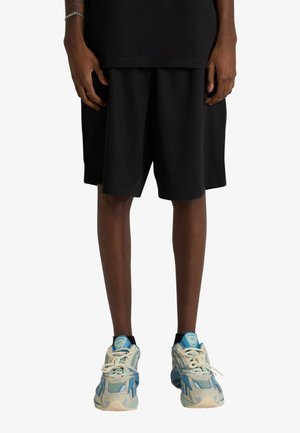 Persona che indossa pantaloncini neri, una maglietta nera e scarpe da ginnastica bianche con dettagli blu, in piedi davanti a uno sfondo bianco uniforme.