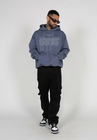 Blå hettegenser med grafisk design, svarte cargo-bukser og svart-hvite sneakers. Modellen bruker solbriller og har et tatovert nakke.