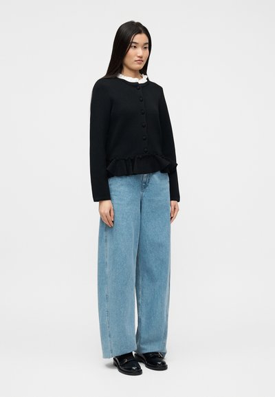 Γυναίκα που στέκεται φορώντας μαύρη ζακέτα με κουμπιά και βολάν στο τελείωμα, ανοιχτό μπλε τζιν με φαρδιά μπατζάκια, μαύρα μοκασίνια και λευκό τοπ με βολάν στο γιακά.