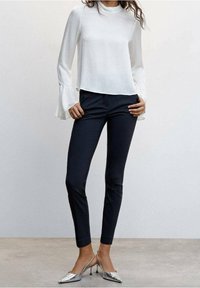 Blouse blanche à manches longues avec poignets cloche, associée à un pantalon bleu marine ajusté et des talons pointus argentés. Tissu lisse et léger.