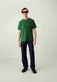 Άνδρας με σκούρα γυαλιά ηλίου, πράσινο κοντομάνικο πουκάμισο, σκούρους παντελόνια με λευκή ραφή και μαύρα παπούτσια, που στέκεται σε ουδέτερο φόντο.