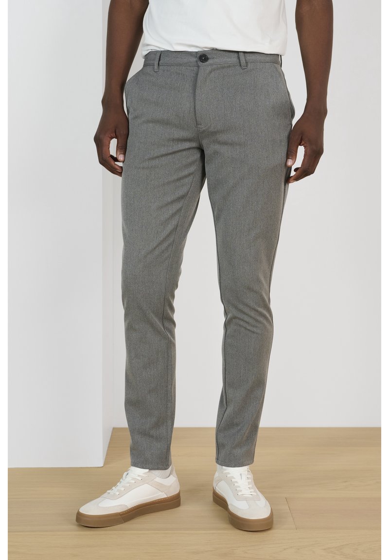 Homme portant un pantalon gris slim et des baskets blanches, debout sur un sol en bois clair contre un mur blanc uni.