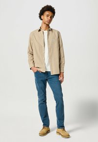 Beige Hemd mit kariertem Muster, kombiniert mit blauen Jeans und beigen Stiefeln. Das Hemd hat einen Kragen und ein gesticktes Logo.