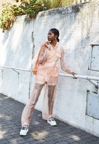 Kvinna klädd i genomskinlig persikofärgad outfit och vita sneakers, lutande mot metallräcke framför en strukturerad vit vägg utomhus.