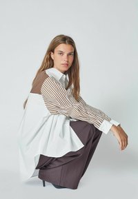 Camicia bianca oversize con maniche a righe marroni, abbinata a pantaloni a gamba larga marroni scuri. Tessuto liscio, polsini con bottoni e colletto.