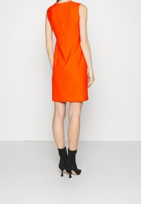 Robe orange sans manches avec des coutures pinces et une fermeture éclair au dos. Portée avec des bottines noires à talon pointu et en matériau texturé.