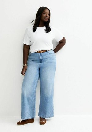 Vrouw met lang zwart haar, gekleed in een wit T-shirt, lichtblauwe wijde jeans met een bruine riem en bruine platte schoenen, staand tegen een witte muur.