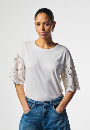 Vrouw met donker haar dat naar achteren is gebonden, draagt een witte top met kanten mouwen en een blauwe spijkerbroek, staat met haar handen in de zakken tegen een effen achtergrond.