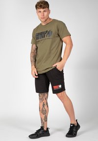 Olivų žalia marškinėliai su "GORILLA WEAR" spauda, derinami su juodais šortais, turinčiais raudoną logotipą. Juodi sportiniai batai ir matomi tatuiruotės ant kojų.