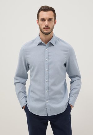 Man draagt lichtblauw overhemd met kraag en donkere broek, staat met handen in zakken tegen een effen achtergrond.