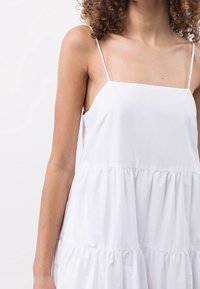 Bílé šaty vyrobené z lehké tkaniny. Mají tenké ramínka, čtvercový výstřih a vrstvený volánový lem. Hladká textura, minimalistický design.