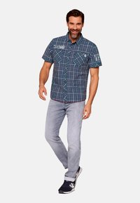 Camicia a quadri blu e verde con maniche corte, due tasche sul petto, abbinata a jeans grigi chiari e sneakers scure.