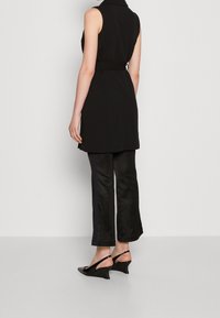 Schwarze ärmellose Weste mit einem Gurt in der Taille, kombiniert mit schwarzen satinartigen, ausgestellten Hosen mit seitlichen Streifen und schwarzen Keil-Slingbacks.