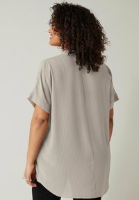 Camisa de manga corta en gris claro, tejido texturizado con patrones sutiles, corte relajado, con aberturas laterales y un dobladillo trasero más largo.