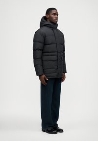 Μαύρο μπουφάν από πούπουλα με κουκούλα, με οριζόντια ραφή, δύο μπροστινές τσέπες και λείες υφή. Φορεμένο με σκούρα τζην και παπούτσια.