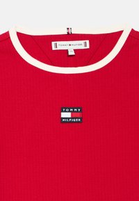 Red ribbed t-shirt with a white collar, featuring a black patch displaying "TOMMY HILFIGER" and a tri-color accent.