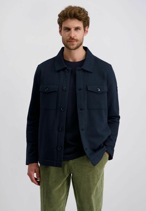 Marineblauwe jas met vier voorzakken, knoopsluiting en gestructureerde kraag, gedragen over een donker shirt en groene ribfluwelen broek.