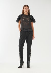 Top in pizzo floreale nero con maniche a volant, abbinato a pantaloni neri con ricami argento sull'orlo e tacchi neri a punta.