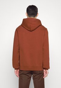 Sweat à capuche marron à coupe décontractée, confectionné dans un tissu doux et texturé. Il comprend une capuche avec cordon de serrage et des coutures minimales dans le dos.