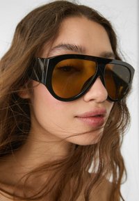 Lunettes de soleil noires surdimensionnées avec des verres teintés ambre et un design à monture épaisse, présentant des surfaces lisses et des bords arrondis.