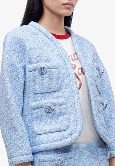 Femme portant un blazer bleu clair texturé et une jupe assortie avec des fermoirs argentés sur une chemise blanche avec col rouge et inscriptions.
