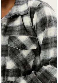 Harmaa ja valkoinen ruudullinen flanellipaita, jossa on napilla suljettava rintatasku ja kauluskallinen kaulus, esittelee pehmeää, teksturoitua kangasta.