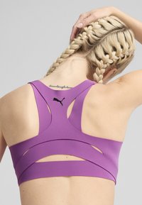 Sujeción deportiva morado con un diseño de espalda nadadora, que presenta detalles recortados y un pequeño logotipo negro en la parte superior de la espalda. Textura suave y material elástico.