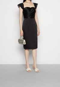 Černé saténové šaty s krátkými vyplněnými rukávy, přiléhavým korzetem a sukní ke kolenům, doplněné stříbrnou kabelkou a béžovými mules na podpatku.