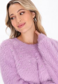 Donna bionda che indossa orecchini a cerchio argentati e un maglione fluffy color lavanda con piccoli strass, che guarda di lato e si tocca il collo.