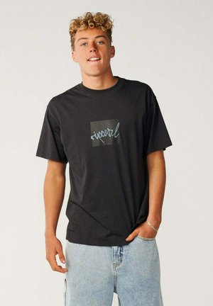 Jeune homme aux cheveux blonds bouclés portant un t-shirt noir Rip Curl et un jean bleu clair, debout avec une main dans la poche.