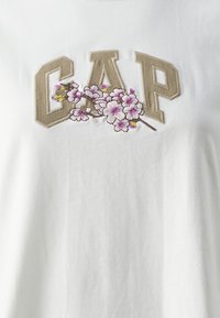 Vit bomullströja med beige broderad "GAP"-logotyp och blommönster i rosa och gult. Slät textur med avslappnad passform.