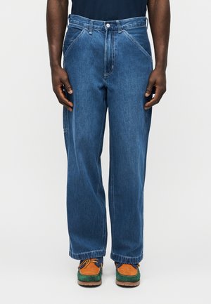 Bărbat purtând blugi albastri lejeri și pantofi cu șiret maro, verzi și albaștri, stând în picioare pe un fundal simplu.