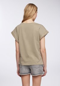 Donna con lunghi capelli biondi indossa una camicia beige a maniche corte con bordo decorativo e pantaloni in denim grigi sfilacciati, in piedi di spalle.