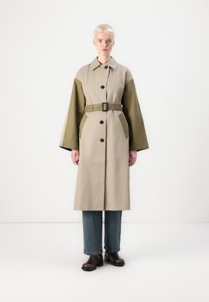 Beige trenchcoat med olivengrønne ærmer, knaplukning, sidelommer og et bredt bælte. Båret over blå denimjeans og brune støvler.