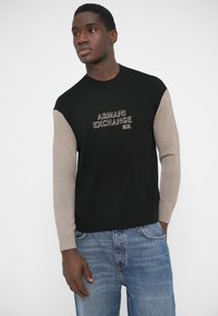 Μαύρο πουλόβερ με μπεζ μανίκια, με κεντημένο το λογότυπο «ARMANI EXCHANGE» σε λευκό. Φορεμένο με ανοιχτόχρωμα μπλε τζιν, με ομοιόμορφη υφή σε όλη την επιφάνεια.