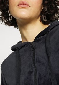Gros plan sur une personne portant un hoodie noir zippé avec des cordons et de grandes boucles d'oreilles en argent géométriques, montrant la partie inférieure du visage et le cou.