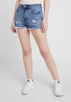 Vrouw draagt high-waisted blauwe denim shorts met rafelige randen en witte sneakers met paars logo, staand tegen een grijze achtergrond.