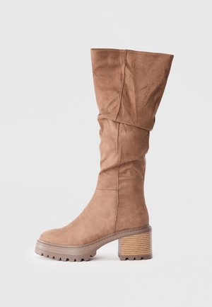 Brązowe zamszowe kozaki do kolan o luźnym kroju, z grubym beżowym obcasem i gumową podeszwą z bieżnikiem zapewniającym stabilność.