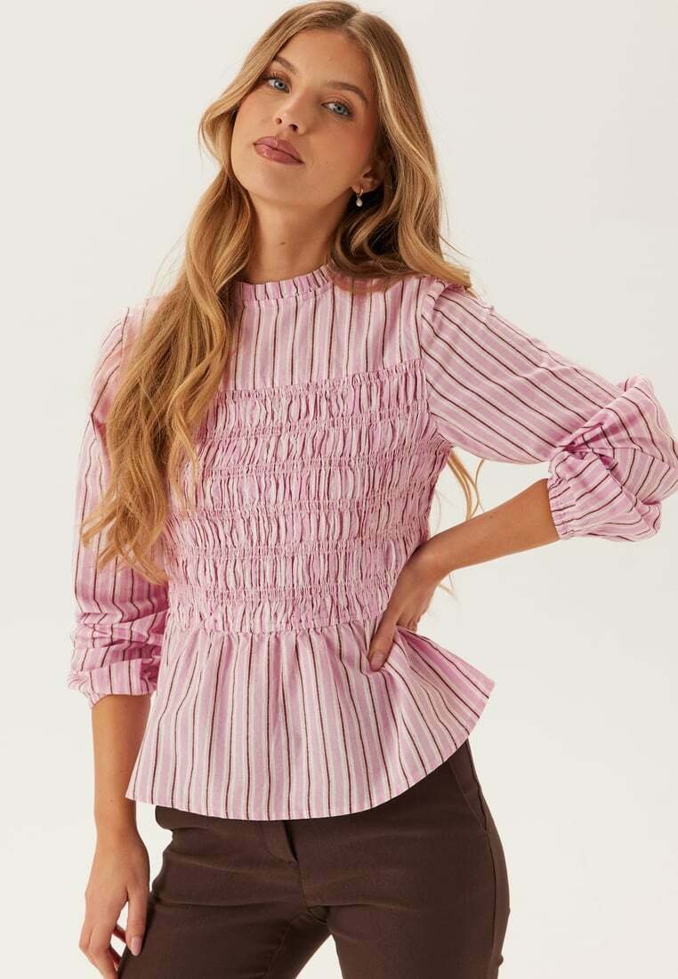 Giovane donna con capelli biondi lunghi indossa una camicetta a righe rosa con corsetto arricciato e maniche a sbuffo, abbinata a pantaloni marrone scuro.