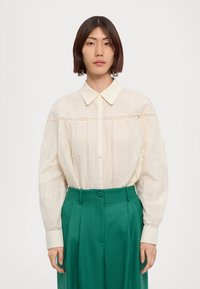White blouse with pleats and lace detailing, paired with high-waisted green trousers. Features a classic collar and button front.