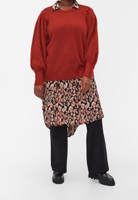 Czerwony sweter z dzianiny z bufkami na tle czarnej sukienki w kwiatowy wzór. Czarne, szerokie spodnie i masywne czarne buty dopełniają strój.