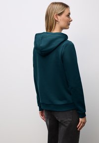 Felpa verde scuro con cappuccio regolabile, polsini e bordino a coste. Il tessuto sembra morbido e leggermente texture. Indossata con pantaloni grigi.